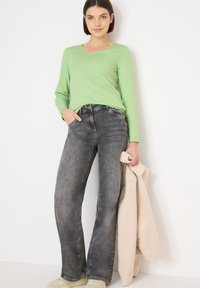 Vaaleanvihreä pitkähihaisen paita yhdistettynä korkeavyötäröisiin harmaisiin farkkuihin. Malli pitää beigeä teksturoitua takkia, poseeraten rennosti ja neutraaleissa kengissä.