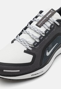 Nike-Laufschuh mit einem weißen Mesh-Obermaterial, grauen Akzenten, strukturierten Details, schwarzer Gummisohle und reflektierenden Schnürsenkeln. AIR ZOOM-Branding sichtbar.