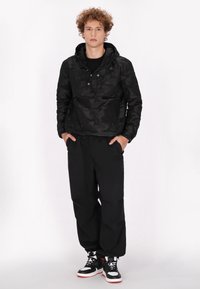 Sort camouflage pulloverjakke med hætte, trykknapper og en lomme; parret med løse sorte bukser og sorte sneakers med røde detaljer.