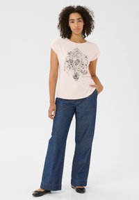Chemise rose clair à manches courtes avec imprimé paisley noir ; associée à un pantalon large en denim bleu foncé et à des chaussures plates noires.