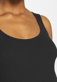 Gros plan sur une personne portant un haut noir sans manches à côtes avec un décolleté en arrondi, montrant l'épaule et le haut de la poitrine.