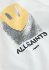 Žlutá abstraktní tvář se šedým stínováním nad textem "ALLSAINTS LONDON" na bílém materiálu.