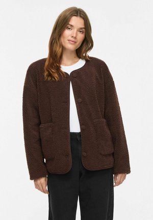 Mujer con chaqueta de forro polar marrón abotonada sobre una camisa blanca y pantalones negros, de pie frente a un fondo blanco liso.