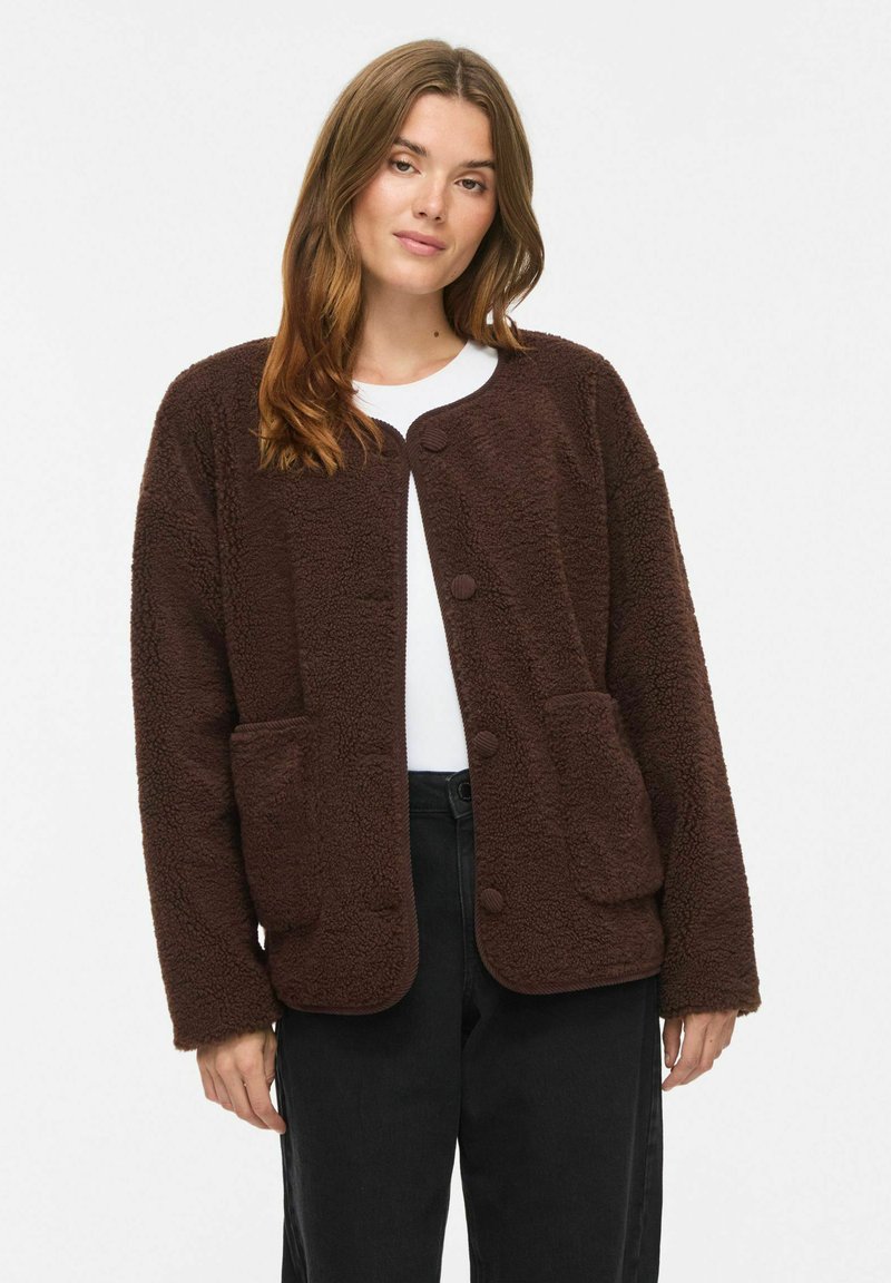 Femme portant une veste en polaire marron boutonnée par-dessus une chemise blanche et un pantalon noir, debout devant un fond blanc uni.