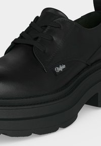 Chaussure noire en cuir à lacets avec semelle plateforme épaisse et petite étiquette de la marque "Buffalo" sur le côté.