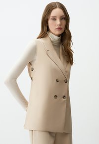 Gilet beige doppiopetto con un design sartoriale, caratterizzato da bottoni visibili e colletto a revers, indossato sopra un leggero dolcevita.