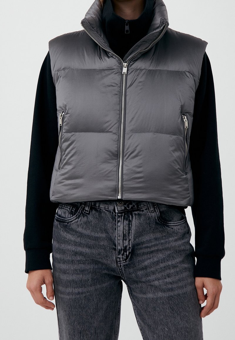 Persona che indossa un gilet imbottito grigio senza maniche sopra una camicia nera a maniche lunghe con jeans grigio scuro, in piedi davanti a uno sfondo semplice.