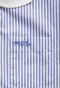 Niebiesko-białe pasiasty materiał z naszywaną kieszenią. Kieszeń ozdobiona haftowanymi inicjałami "RSL" w jasnoniebieskiej nici. Gładka tekstura.