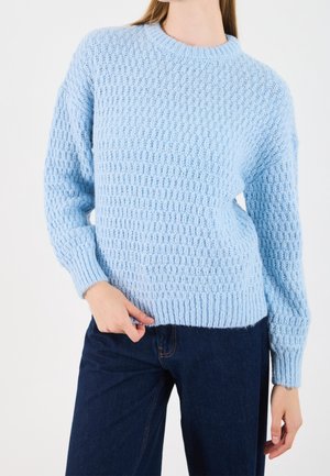 Femme portant un pull en maille texturé bleu clair à manches longues et un jean bleu foncé, posant avec une main près de sa poche.