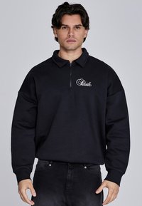 Sudadera negra con cuello con cremallera, ajuste holgado y logo en el lado izquierdo del pecho. Fabricada con material suave, con mangas texturizadas y de color sólido.