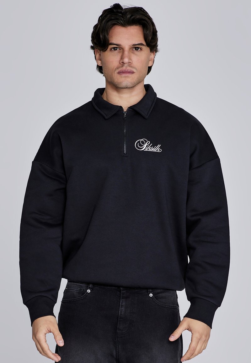 Sudadera negra con cuello con cremallera, ajuste holgado y logo en el lado izquierdo del pecho. Fabricada con material suave, con mangas texturizadas y de color sólido.