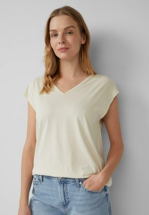Frau mit hellbraunem Haar in einem cremefarbenen kurzärmligen V-Ausschnitt-Oberteil und hellblauen Jeans, die mit einer Hand in der Tasche an einer grauen Wand steht.