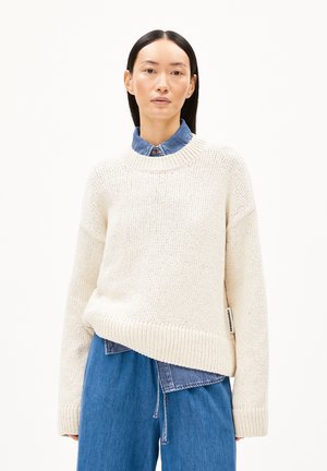 Cream-colored knitted sweater with a wide neckline and ribbed cuffs, layered over a chambray shirt, featuring a slightly oversized fit.