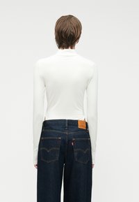 Λευκή μπλούζα με μακριά μανίκια κ�αι ψηλό γιακά, συνδυασμένη με σκούρους τζιν μπλε παντελόνες που διαθέτουν πίσω τσέπες και καφέ δερμάτινη ετικέτα στη μέση.