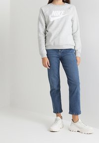 Sudadera gris con logo blanco de Nike, combinada con jeans azules y zapatillas blancas. Presenta un cuello redondo y puños acanalados.