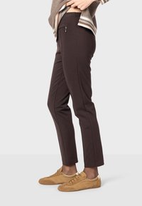 Braune Slim-Fit-Hosen aus dehnbarem Material, mit einer seitlichen Tasche mit Reißverschluss. Kombiniert mit hellbraunen Sportschuhen.