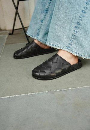 Persona con zapatos negros de tela trenzada y jeans de pierna ancha de color azul claro con dobladillo deshilachado, de pie sobre un suelo de baldosas grises junto a una pata de silla.