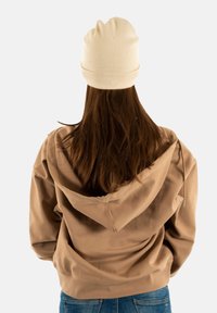 Persona con lunghi capelli castani che indossa un berretto beige, una felpa color sabbia e jeans blu, vista di spalle su uno sfondo neutro.