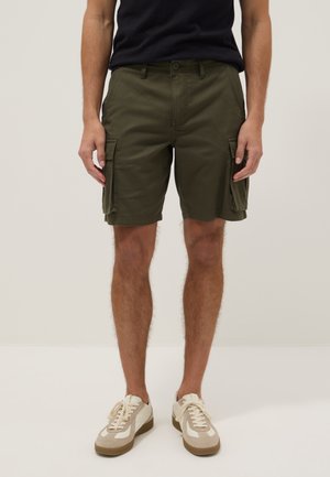 Uomo che indossa pantaloncini cargo verde oliva con tasche laterali, maglia nera e sneakers beige su uno sfondo chiaro uniforme.