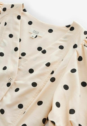 Helle beige Bluse mit schwarzen Pünktchen, V-Ausschnitt, Knopfverschluss und puffigen Ärmeln, aus einem Leinenmix gefertigt.