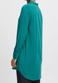 Chemise à manches longues couleur sarcelle avec un col, fabriquée en tissu lisse. Comprend des poignets boutonnés et un ourlet courbé qui s'étend en dessous des hanches.