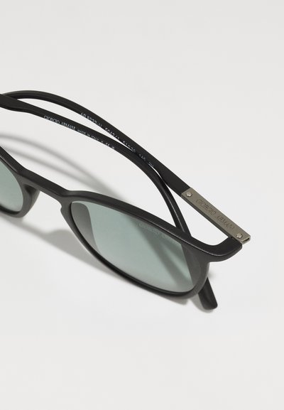 Black sunglasses with rounded lenses, green tinted glass, matte finish, and silver logo accent on the arms. Sleek, modern design.