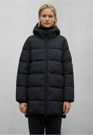 Kvinna iklädd en svart vadderad vinterjacka med hög krage och huva, stående mot en enfärgad grå bakgrund.