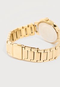 Λουράκι ρολογιού από χρυσό μέταλλο με κλείσιμο που διπλώνει, εμφανιζόμενο από πίσω πάνω σε ανοιχτογκρι επιφάνεια.