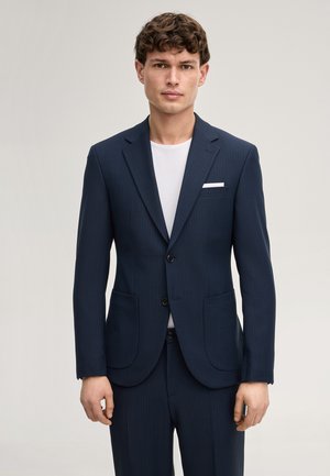 Junger Mann trägt einen marineblauen Nadelstreifenanzug mit zwei Knöpfen, aufgesetzten Taschen und einem weißen Einstecktuch über einem weißen Rundhalsausschnitt-Shirt.