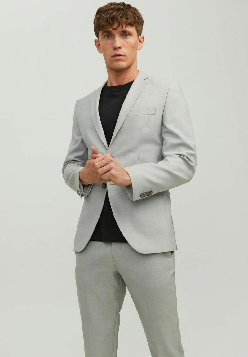 Light gray suit made of textured fabric, featuring a single-button jacket with a pocket and matching tailored trousers over a black shirt.