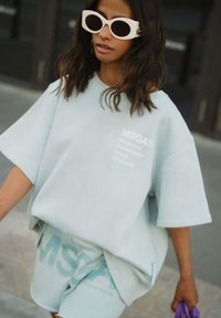 Overmaat mintgroene sweatshirt en shorts, beide met witte "MSGA" branding. Heeft een zachte textuur en een losse fit, gecombineerd met witte zonnebril.