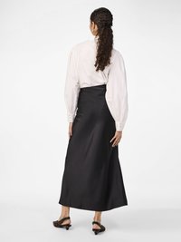 Mulher com cabelo encaracolado, vestindo uma blusa branca e uma saia midi preta de cintura alta, usando sapatos de salto baixo de alça na cor leopardo, vista de costas.