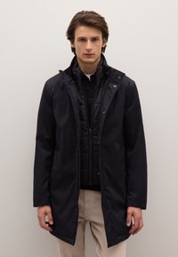 Abrigo impermeable negro con capucha, con cierre frontal de cremallera y broches a presión. Llevado sobre un chaleco acolchado negro, combinado con pantalones beige.