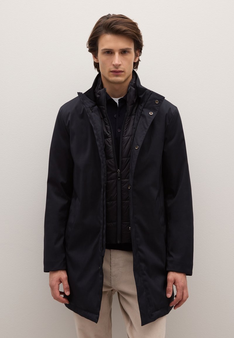 Abrigo impermeable negro con capucha, con cierre frontal de cremallera y broches a presión. Llevado sobre un chaleco acolchado negro, combinado con pantalones beige.