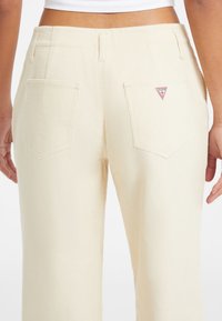 Vista posteriore di una persona che indossa pantaloni in velluto a coste color crema con due tasche posteriori, una delle quali presenta un logo Guess rosso e bianco.