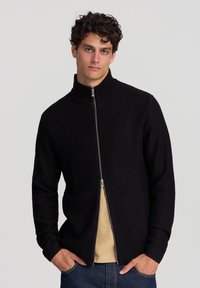 Uomo con capelli ricci che indossa un maglione nero a zip parzialmente chiuso sopra una camicia beige e jeans blu, in piedi davanti a uno sfondo neutro.