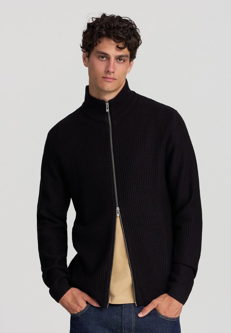 Uomo con capelli ricci che indossa un maglione nero a zip parzialmente chiuso sopra una camicia beige e jeans blu, in piedi davanti a uno sfondo neutro.