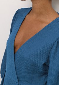 Robe texturée bleue avec un décolleté en V profond et de subtils plis aux épaules, mettant en valeur une silhouette structurée mais douce.