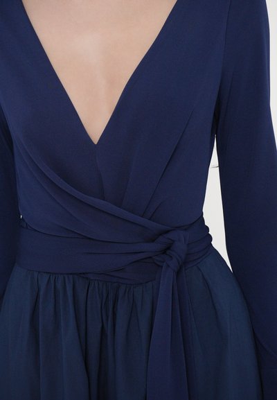 Tmavě modré šaty s hlubokým výstřihem do V, dlouhými rukávy a páskem v pase. Tkanina je hladká s plynulou, plisovanou sukní.