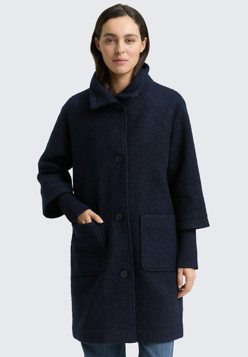 Marineblauer Mantel aus einem strukturierten, wollartigen Stoff, mit breitem Kragen, Knopfvorn und zwei Fronttaschen. Der Mantel hat Dreiviertelärmel.