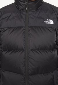 Czarna kurtka puchowa z wysokim kołnierzem, widocznymi szwami i zamkiem błyskawicznym z przodu. Posiada białe logo "The North Face" po lewej stronie.