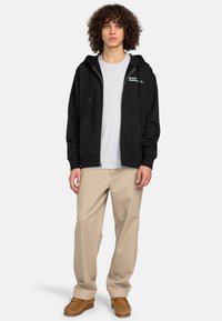 Black zip-up hoodie with kangaroo pockets and drawstring hood, worn over a light gray shirt. Paired with light beige pants and brown shoes.