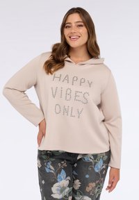 Hanorac bej deschis dintr-un material moale, având inscripția "HAPPY VIBES ONLY" din pietricele argintii. Asortat cu pantaloni înfășurați cu un model floral întunecat.