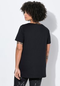 Camiseta negra de manga corta con escote redondo, dobladillo trasero ligeramente más largo y tela de algodón suave. Se lleva con pantalones negros estampados.
