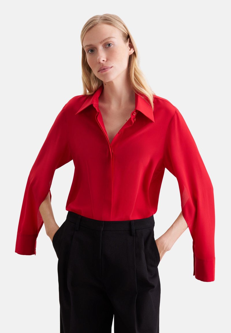Femme aux longs cheveux blonds porte un chemisier rouge vif à manches ouvertes et un pantalon noir taille haute, debout les mains dans les poches.