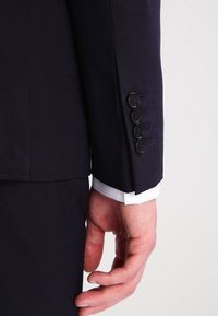 Marinblå kostymjacka med ärm som har fyra svarta knappar på en strukturerad tyg, som avslöjar en vit skjortärm nedanför.