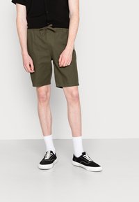 Shorts vert olive avec une ceinture élastique et un cordon de serrage, fabriqués en tissu. Portés avec des baskets noires et des chaussettes blanches.