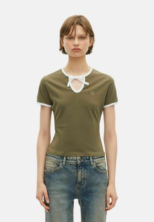 Jonge vrouw in een olijfgroene shirt met korte mouwen en witte bies en strikje bij de hals, gecombineerd met vervaagde blauwe jeans, staand tegen een witte achtergrond.