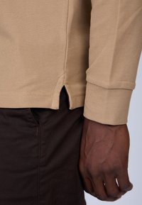 Chemise en tricot beige à manches longues avec un tissu texturé, présentant une fente sur le côté à l'ourlet et une manche à poignet. Associée à un pantalon marron foncé.