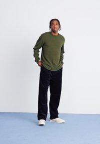 Pull à manches longues vert olive, pantalon sombre ample et baskets blanches. Texture lisse, design décontracté, debout devant un fond clair.